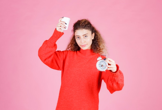 woman holding an alarm clock and a cup of coffee pointing at her morning routine.