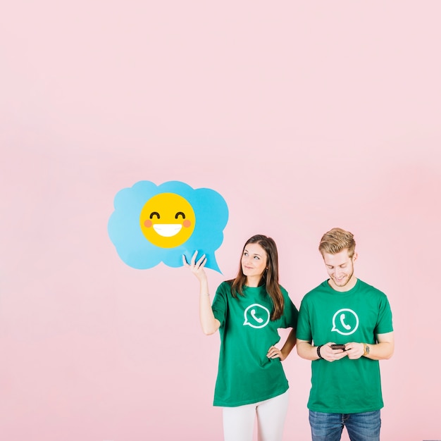 Woman holding blue speech bubble with laughing emoji near man using cellphone