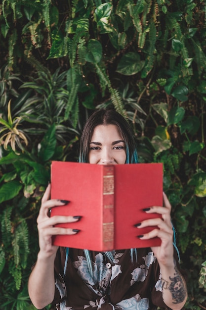 Woman holding book in front of her mouth standing against plant