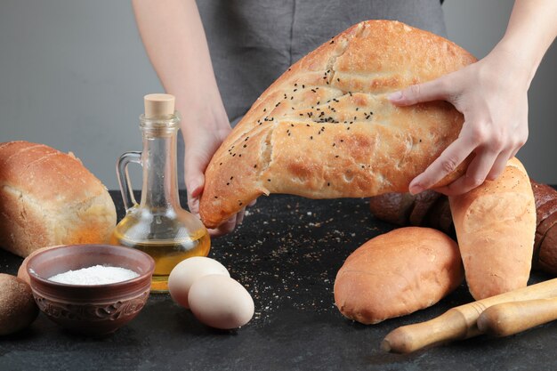 Woman holding bread on dark table with eggs, flour bowl and glass of oil.