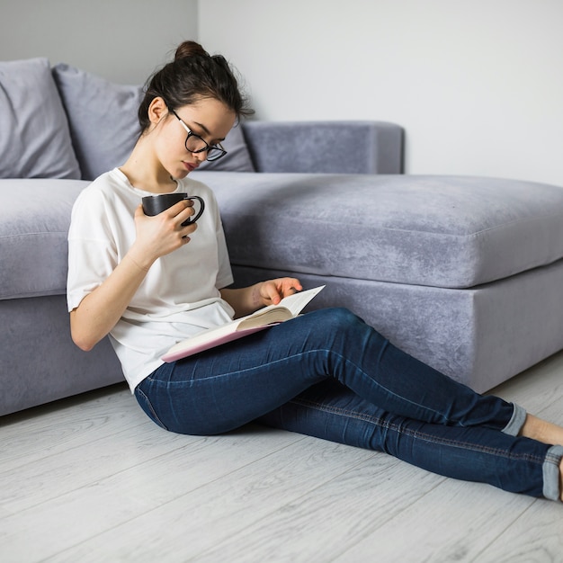 Woman holding cup and reading book on floor