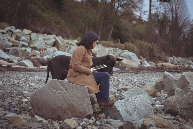 Woman holding digital tablet by dog at beach