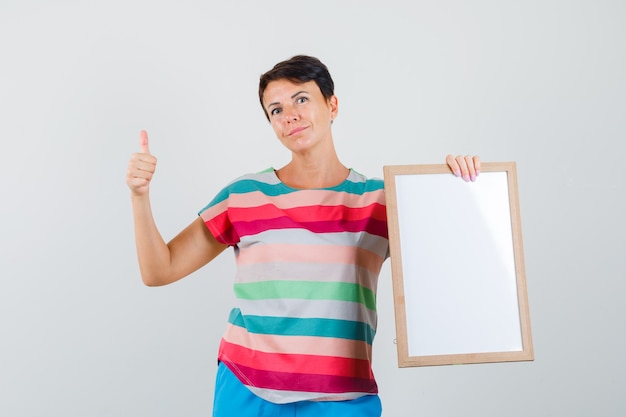 Женщина держит пустую рамку, показывая большой палец вверх в полосатой футболке, штанах и выглядит уверенно.