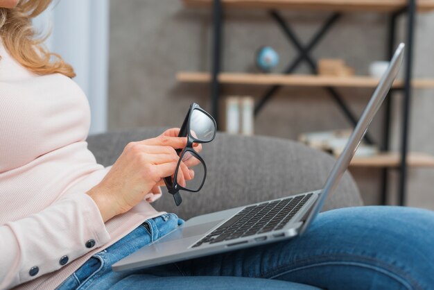 Woman holding eyeglasses in hand sitting on sofa with an open laptop on her lap