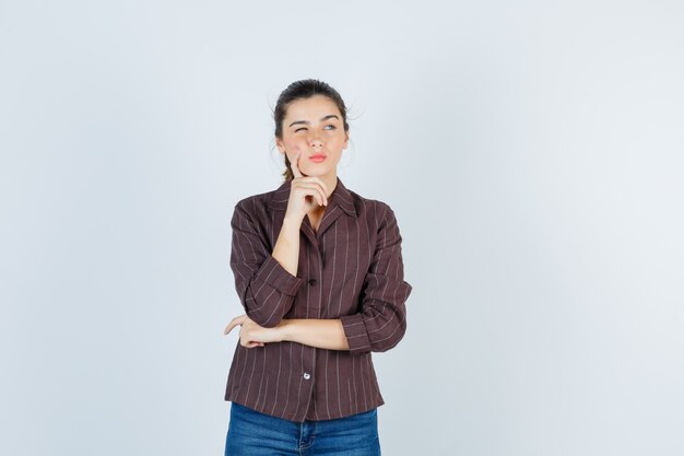 woman holding finger on cheek, looking away in brown striped shirt and looking thoughtful. front view.