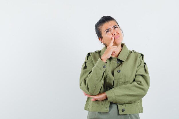 Woman holding finger near mouth in jacket, t-shirt and looking hesitant