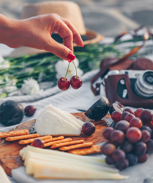 woman holding fruit with cheese and fruits on wooden cutting board and camera, hat and flowers in beach.