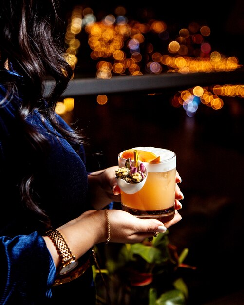 Woman holding a glass of orange cocktail looking from the night city view
