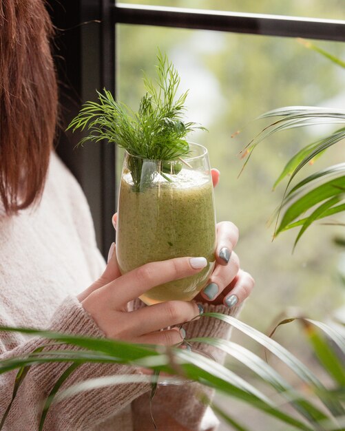 Woman holding a green detox smoothie with dills by the window.