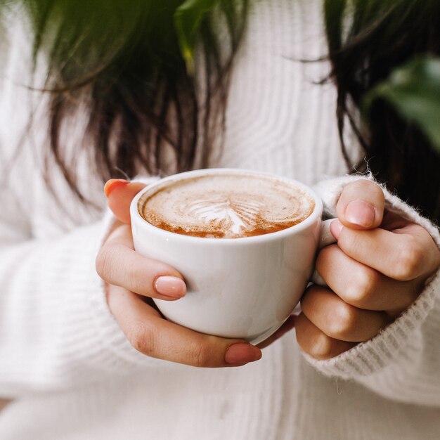 白いニットジャケットでホットコーヒーカップを保持している女性