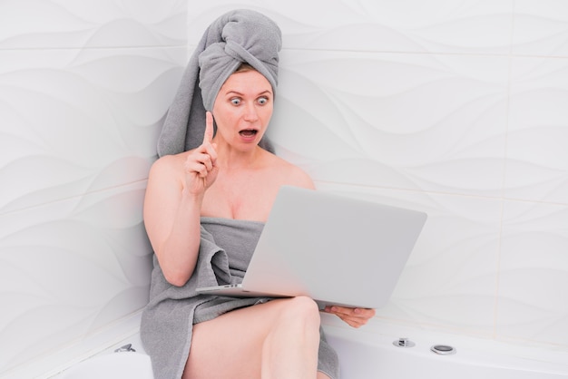 Женщина держит ноутбук в ванне и смотрит в изумлении