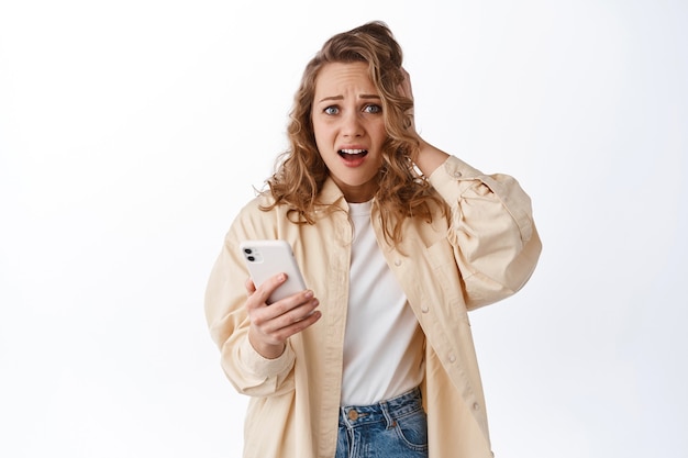Free photo woman holding phone and look in panic, feeling anxious and worried about something posted online, standing with smartphone against white wall