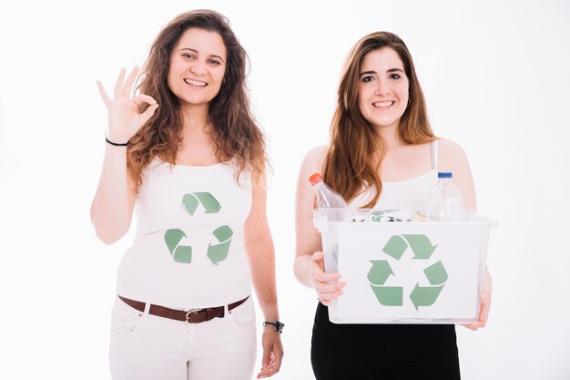 Woman holding recycle bin filled with trash and her friend showing ok sign