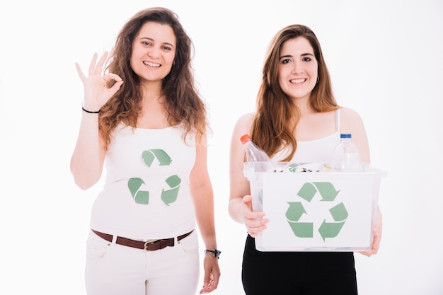 Woman holding recycle bin filled with trash and her friend showing ok sign