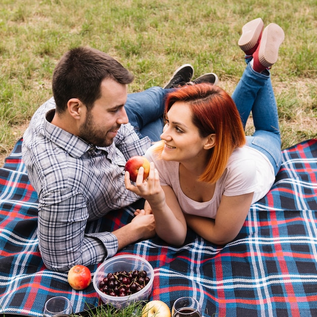 Woman holding red apple in hand lying with her boyfriend on blanket