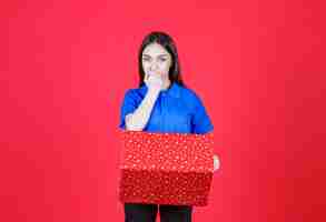 Free photo woman holding a red gift box with white dots on it and looks confused and hesitating.