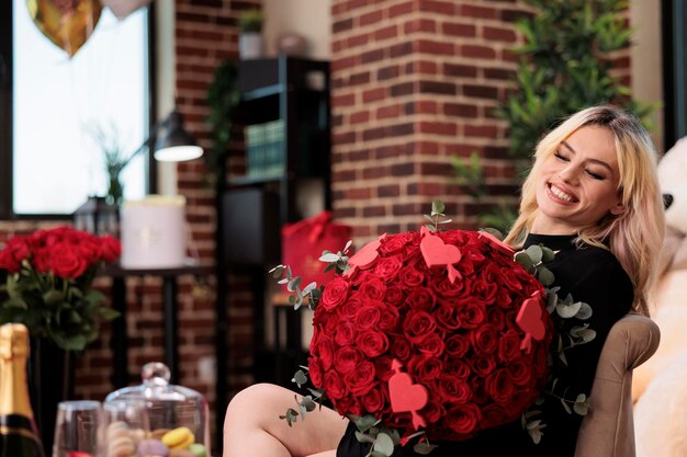 Woman holding red roses bouquet with hearts decoration, laughing in room filled with luxury presents, valentines day romantic date. Excited blonde girl with expensive flowers sitting