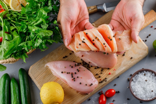 Woman holding sliced chicken breast with greens, cucumber, lemon, salt on a cutting board on gray surface, top view