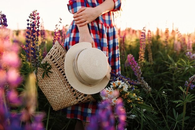 夕暮れの花畑に立っているわらバッグと帽子を持っている女性。