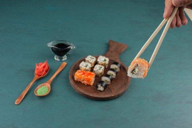 Woman holding sushi roll with chopsticks on blue table with pickled ginger and soy sauce.