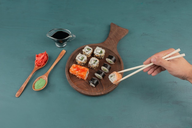 Woman holding sushi roll with chopsticks on blue table with pickled ginger and soy sauce.