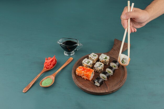 Woman holding sushi roll with chopsticks on blue table with pickled ginger and soy sauce.