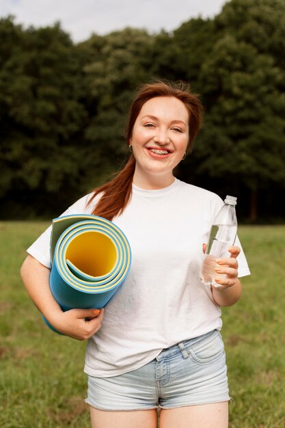 Женщина, держащая бутылку с водой и коврик для йоги на открытом воздухе