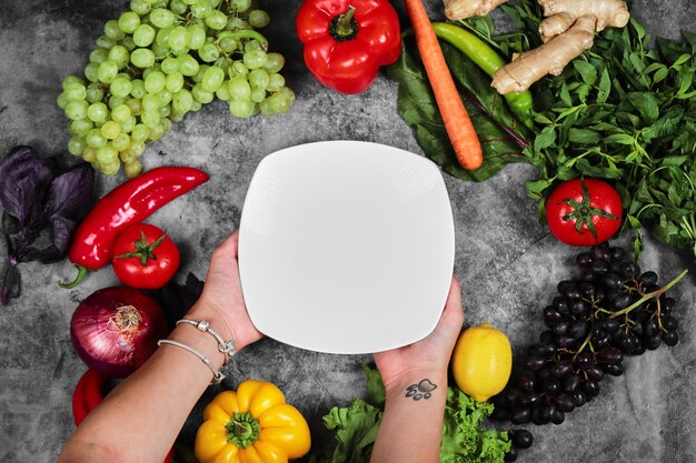 Woman holding white plate on marble background with fresh vegetables. 