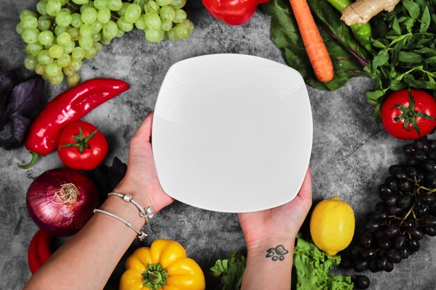 Woman holding white plate on marble background with fresh vegetables. 