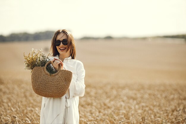 밀 밭에서 산책 하는 밀 짚 가방에 야생화 꽃다발을 들고 여자. 흰 옷을 입고 여름 들판을 걷는 갈색 머리 여자