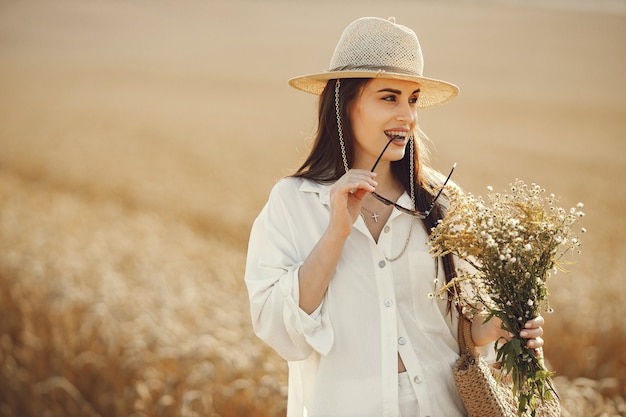 麦畑を歩いて、わらの袋に野花の花束を持っている女性。白い服を着て夏のフィールドを歩くブルネットの女性