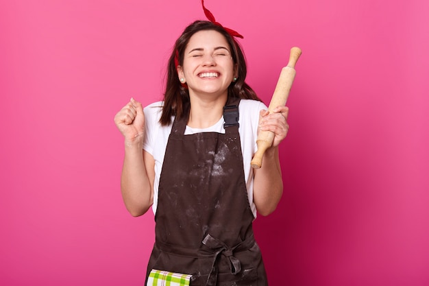 Woman holds baking rolling pin, wears brown apron, white t shirt.