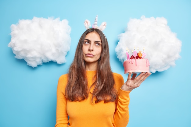 woman holds birthday cake with burning candles thinks about party and celebration dressed in orange jumper isolated on blue 