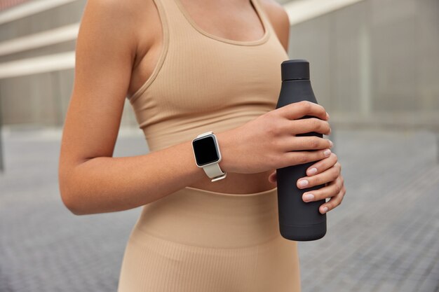 woman holds bottle of protein drink or water has regular training outdoors to keep fit dressed in tracksuit uses smartwatch to monitor fitness activity