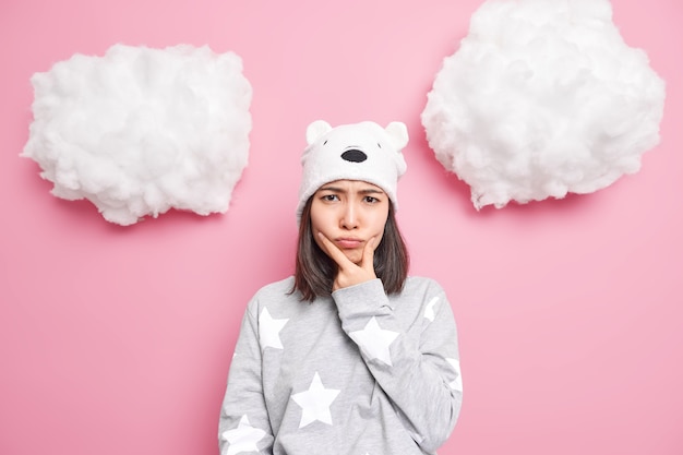 Free photo woman holds chin and looks offended feels angry awakes in bad mood dressed in slumber suit soft hat with bear ears isolated on pink