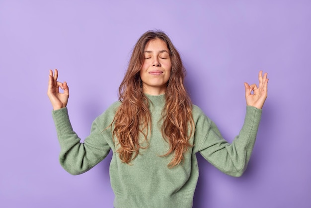 Woman holds hands in zen om gesture keeps eyes closed meditates indoor dressed casually keeps emotions under control peace and patience breathes air freely isolated on purple wall Free Photo