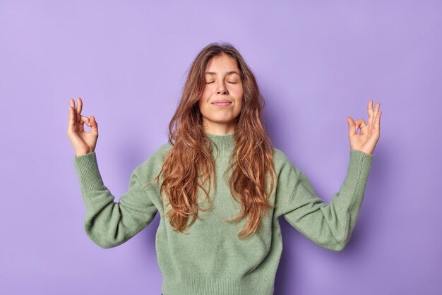 女性は禅のジェスチャーで手を握り、目を閉じて瞑想し、カジュアルな服装で感情をコントロールし、忍耐力は紫色の壁に自由に隔離された空気を吸います