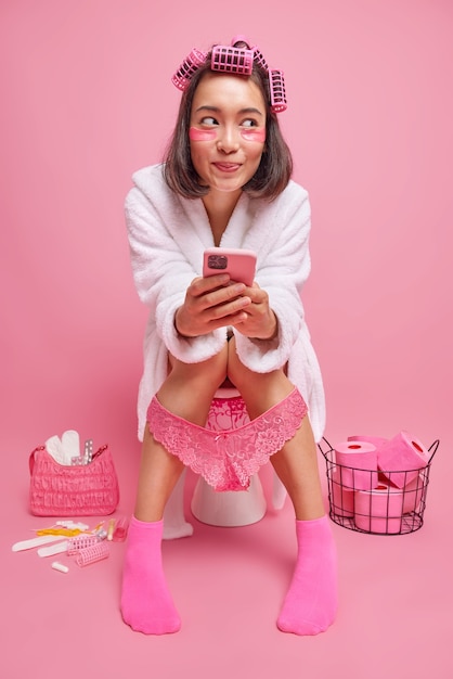 woman holds mobile phone in hands sits on toilet bowl has thoughtful expression looks away makes hairstyle with rollers applies beauty patches isolated on pink wall in bathroom