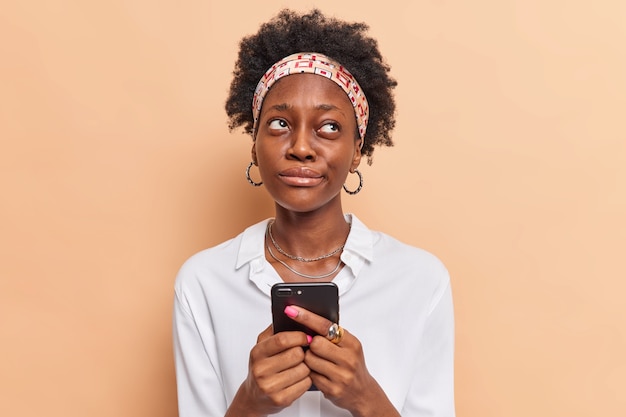 La donna tiene il telefono cellulare riflette su come rispondere alla domanda sotto il post sul sito web ricorda il nome del prodotto prima di navigare in internet vestita con abiti eleganti beige
