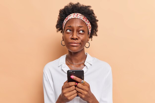  woman holds mobile phone ponders how to answer question under post on website remembers name of product before browsing internet dressed in stylish clothes beige 