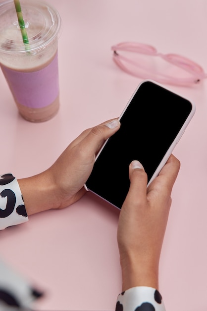 woman holds mobile phone with empty screen sits at pink table with milkshake sunglasses around browses website information