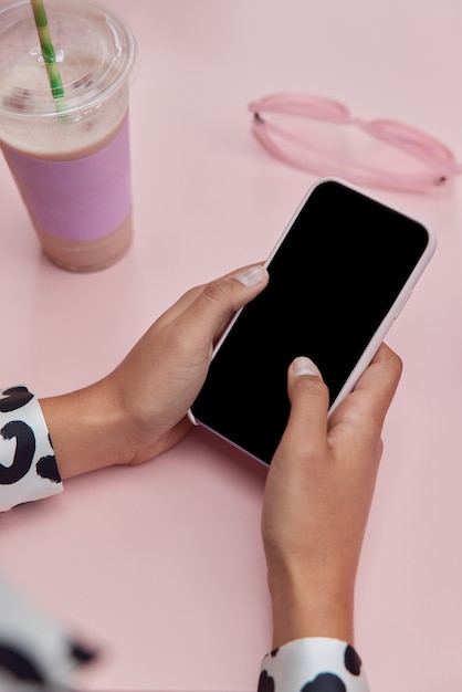 женщина держит мобильный телефон с пустым экраном сидит за розовым столом в солнцезащитных очках с молочным коктейлем и просматривает информацию на веб-сайте