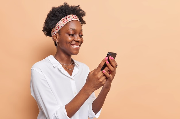 Free photo woman holds modern mobile phone surfs net checks newsfeed in social networks wears headband white shirt isolated on beige