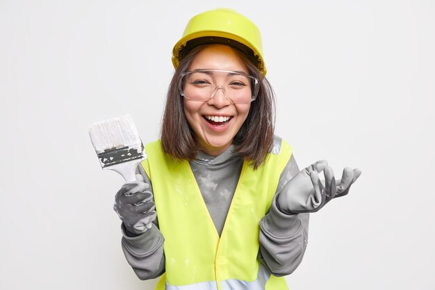 woman holds paint brush remodels house wears safety equipment and uniform smiles happily 