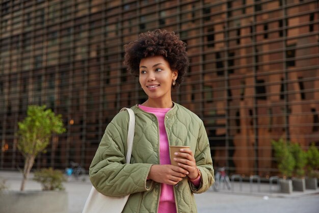 woman holds paper cup of coffee spends leisure time outdoors wears jacket carries bag enjoys recreation dreams about something