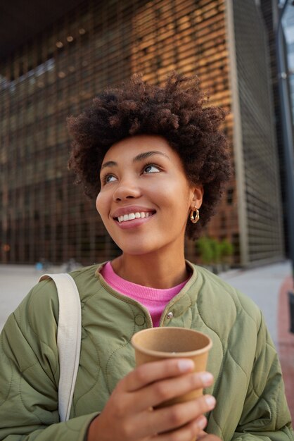 woman holds paper cup of hot beverage looks away with glad expression smiles toothily walks in city dressed in jacket enjoys favorite drink while having promenade