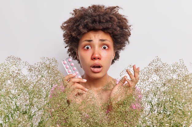 woman holds pills to cure allergy surrounded by bouquet of wildflowers has red swollen eyes stands on white 