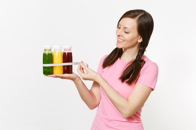 여자는 병에 녹색, 빨간색, 노란색 해독 스무디를 들고 흰색 배경에 분리된 테이프를 측정합니다. 적절한 영양, 채식 음료, 건강한 생활 방식, 다이어트 개념. 복사 공간, 플랙.