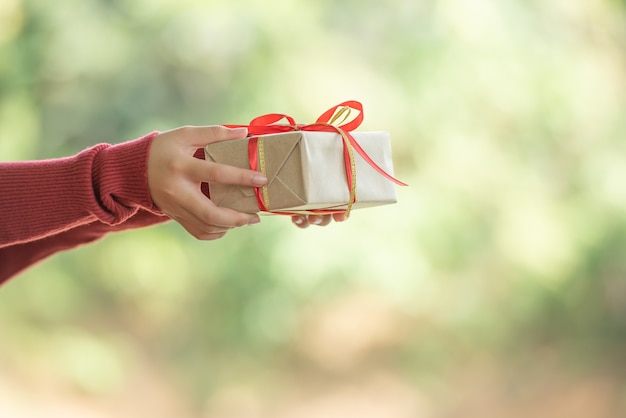 A woman holds a small gift box in beautiful hands. The girl is outdoors against the backdrop green leaves bokeh out of focus background from nature forest.