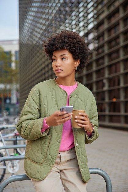 女性はスマートフォンとコーヒーと紙コップを持って人生を楽しんでいます都市の設定を見て休暇旅行で再現しますブログのための良い一日の使用アプリを楽しんでいます